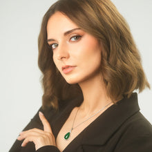 Woman wearing a black blazer with a green pendant necklace against a plain background